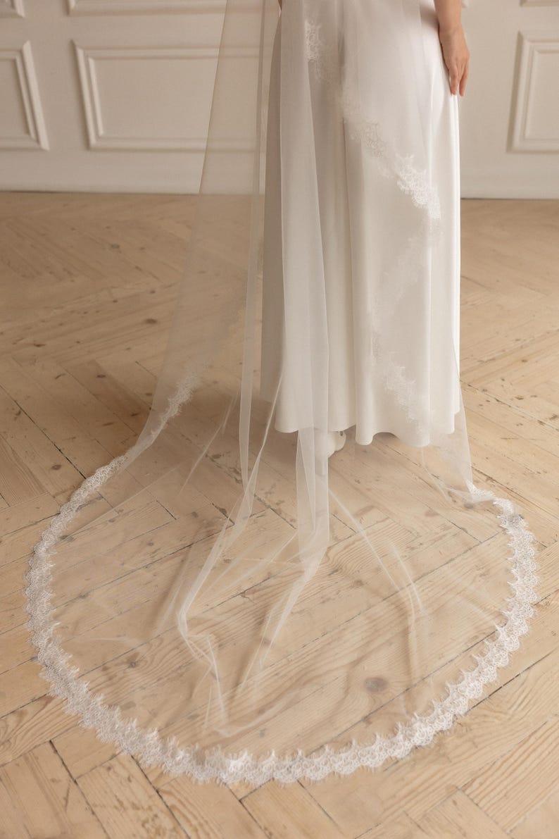 May include: A long, sheer white wedding veil with a delicate lace trim. The veil is draped over a white dress, creating a flowing, elegant look. The floor is made of wooden planks.