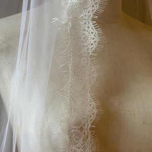 Pu&ograve; includere: Un velo da sposa bianco trasparente con un delicato bordo in pizzo. Il pizzo presenta un intricato motivo floreale lungo il bordo, aggiungendo un tocco di eleganza. Il velo &egrave; drappeggiato su un manichino, mostrando il suo tessuto fluido e il suo design.