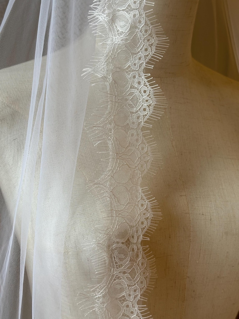 Pu&ograve; includere: Un velo da sposa bianco trasparente con un delicato bordo in pizzo. Il pizzo presenta un intricato motivo floreale lungo il bordo, aggiungendo un tocco di eleganza. Il velo &egrave; drappeggiato su un manichino, mostrando il suo tessuto fluido e il suo design.