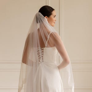 Puede incluir: Una novia con un vestido de novia blanco y velo. El vestido tiene espalda de cors&eacute; y tirantes finos. El velo transparente tiene un ribete de encaje. La novia est&aacute; de lado, mirando hacia otro lado.