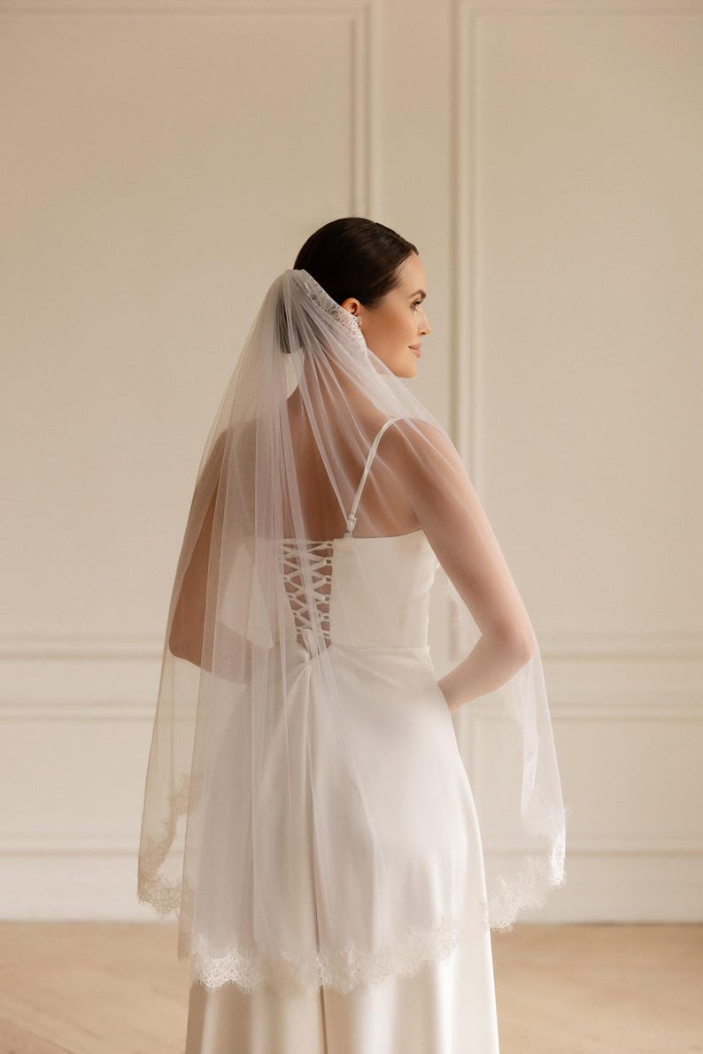 Puede incluir: Una novia con un vestido de novia blanco y velo. El vestido tiene espalda de cors&eacute; y tirantes finos. El velo transparente tiene un ribete de encaje. La novia est&aacute; de lado, mirando hacia otro lado.