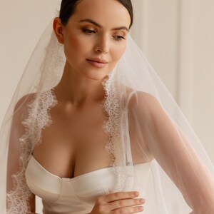 Puede incluir: Una novia con un vestido de novia de sat&eacute;n blanco y un velo transparente con delicados bordes de encaje. El velo enmarca la cara y los hombros, creando un aspecto suave y rom&aacute;ntico. El vestido tiene un corpi&ntilde;o ajustado y un dise&ntilde;o simple y elegante.