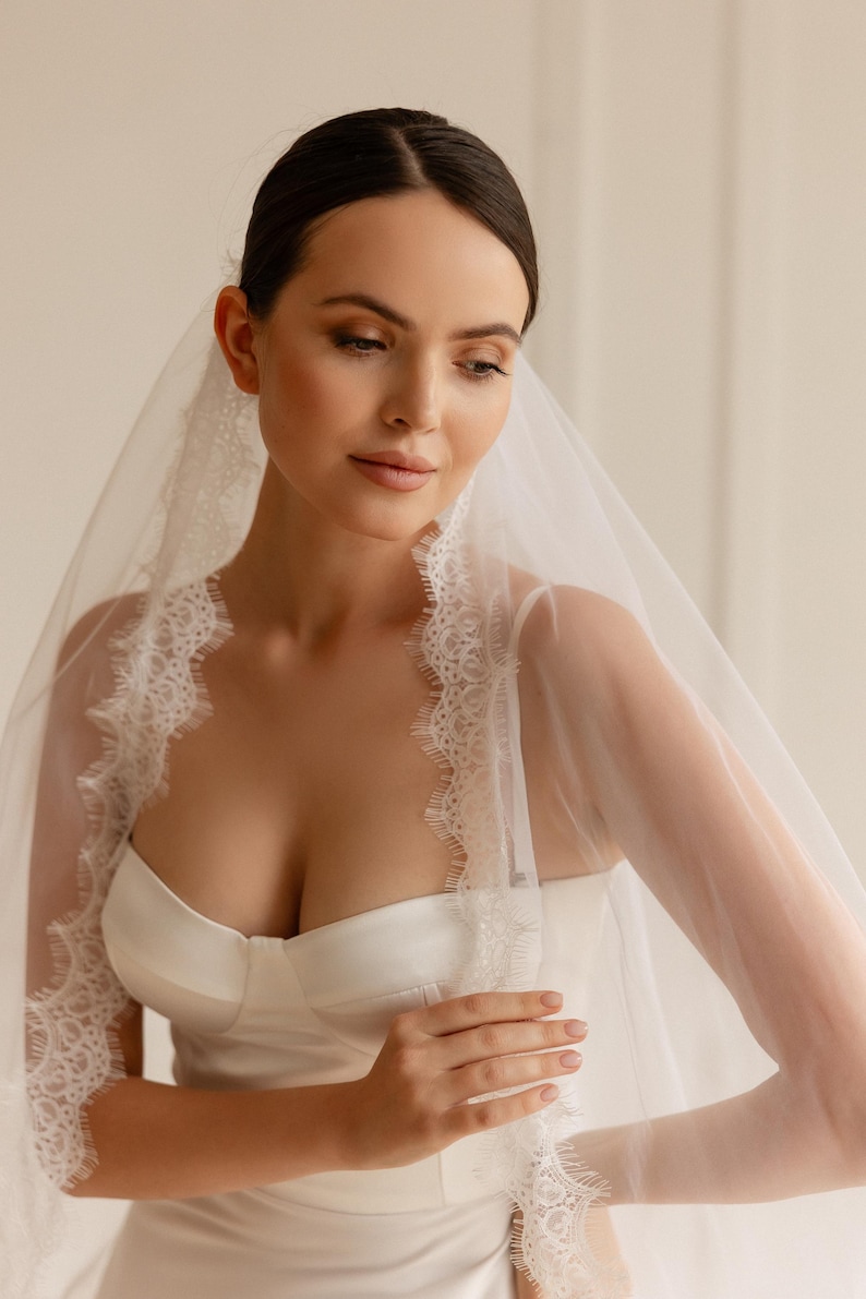 Puede incluir: Una novia con un vestido de novia de sat&eacute;n blanco y un velo transparente con delicados bordes de encaje. El velo enmarca la cara y los hombros, creando un aspecto suave y rom&aacute;ntico. El vestido tiene un corpi&ntilde;o ajustado y un dise&ntilde;o simple y elegante.