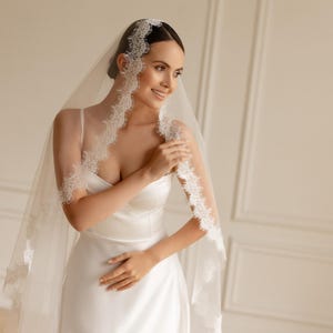 May include: A bride wearing a white satin wedding dress and a sheer veil with delicate lace trim. The veil is draped over the head and shoulders, creating a soft, romantic look. The bride is smiling, and the background is a neutral color.