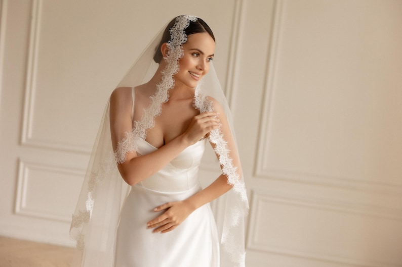 May include: A bride wearing a white satin wedding dress and a sheer veil with delicate lace trim. The veil is draped over the head and shoulders, creating a soft, romantic look. The bride is smiling, and the background is a neutral color.
