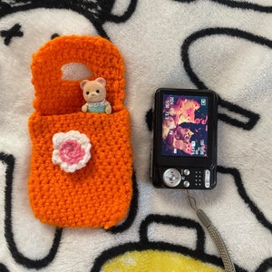 May include: A small orange crocheted bag with a white flower and a pink center. A small brown bear wearing a blue dress is sitting inside the bag. A black digital camera is next to the bag.