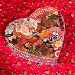 May include: A heart-shaped tin box with a collage of images and stickers, including a Ty Beanie Baby, a Hello Kitty sticker, and a crayon. The box is pink with a scalloped edge and the word "Quebec" is visible.