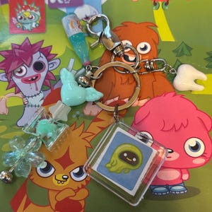 May include: A collection of colorful keychains and charms featuring cartoon characters and novelty designs. Includes a keychain with a bottle charm, a bird charm, a tooth charm, and a clear square charm with a cartoon character.
