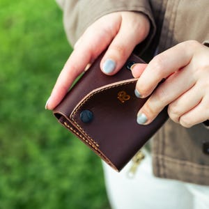 以下が含まれることがあります： カーブしたフラップとスナップ留めが付いた、濃い茶色の革製財布。財布にはステッチされた縁と金色のエンボス加工のデザインがあります。背景はぼやけており、緑の草と茶色のジャケットを着た人がいます。