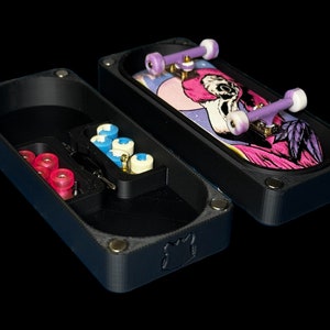 May include: A black fingerboard case with a fingerboard inside. The case has a compartment for storing wheels and other accessories. The fingerboard has a pink and purple graphic with a skull design.
