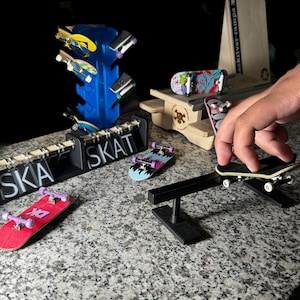 May include: A collection of fingerboard skateboards and ramps, including a black ramp with a skateboard on it, a wooden ramp with a skull design, and a blue and black fingerboard rack with the word "SKA" written on it.