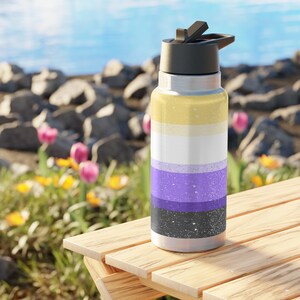 Pode incluir: Uma garrafa de água de aço inoxidável com tampa preta e um design da bandeira do orgulho não binário. A bandeira apresenta listras horizontais em amarelo, branco, roxo e preto. A garrafa está em uma mesa de madeira.