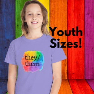 May include: A purple youth-sized t-shirt with a rainbow watercolor design and the text "they/them" in black.