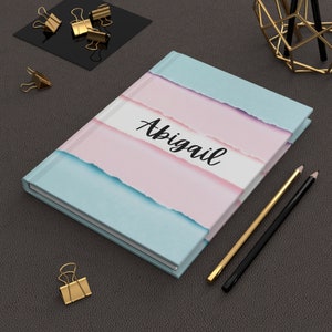 May include: A blue and pink notebook with a ripped paper design and the name "Abigail" written in black on the front cover.