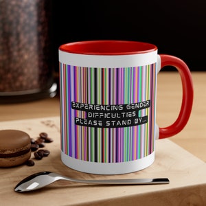 May include: White ceramic mug with a red rim and handle. The mug has a colorful striped pattern and the text "Experiencing gender difficulties please stand by..." printed on the front.
