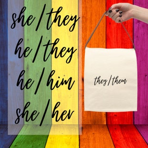 May include: A canvas tote bag with a brown strap is held against a rainbow-colored wooden background. The bag displays the text "they/them" in black script. The background features the phrases "she/they", "he/they", "he/him", and "she/her".