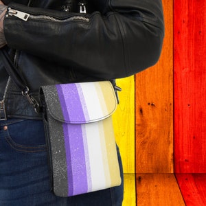 May include: A black leather crossbody bag with a zipper closure. The bag has a white, yellow, purple, and black striped pattern. The bag is worn by a person wearing a black leather jacket and blue jeans.