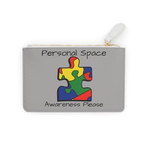 Könnte beinhalten: Graue Reißverschlusstasche mit farbenfrohem Puzzleteil-Design. Die Texte "Personal Space" und "Awareness Please" sind auf der Tasche aufgedruckt. Der Reißverschlussanhänger ist weiß.