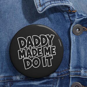 Könnte beinhalten: Ein runder Ansteckbutton mit schwarzem Hintergrund und weißem Text, der DADDY MADE ME DO IT in einer fetten serifenlosen Schrift zeigt. Der Button ist an einem blauen Jeansstoff mit sichtbaren Nähten und einem dunklen Metallknopf befestigt.