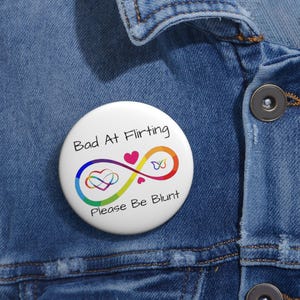 May include: A round white pin-back button featuring a rainbow-colored infinity symbol, a small pink heart, and a butterfly outline. The text reads Bad At Flirting and Please Be Blunt in black lettering. The button is attached to a blue denim fabric.