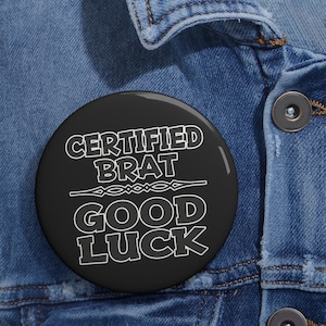 May include: A round black pin-back button featuring white text that reads CERTIFIED BRAT GOOD LUCK with a decorative horizontal line separating the two phrases. The button is attached to a blue denim fabric surface with visible metal buttons.