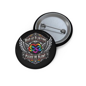 May include: A round pin-back button featuring a black background with a metallic emblem. The design includes a rainbow-colored infinity heart symbol, silver wings, and the text "BAD AT FLIRTING" and "PLEASE BE BLUNT" in a bold, silver font.