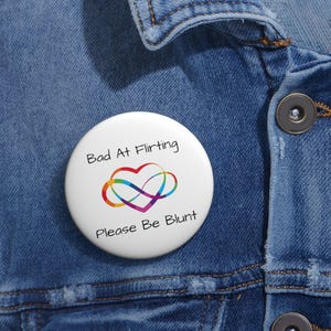 May include: A circular white pinback button featuring a colorful infinity heart symbol in the center. The text "Bad At Flirting" is printed above the symbol, and the text "Please Be Blunt" is printed below the symbol in black lettering.