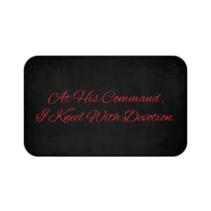 May include: A rectangular black bath mat with rounded corners featuring the red text "At His Command, I Kneel With Devotion." written in a cursive script font centered on the surface of the mat.