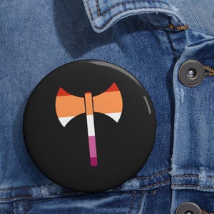 May include: A black button with a graphic of a double-bladed axe in the colors of the lesbian pride flag: orange, white, and pink. The axe is centered on the button, which is set against a blue denim background.
