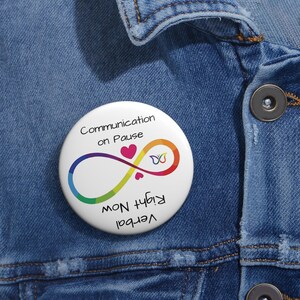 Könnte beinhalten: Ein runder weißer Ansteckbutton mit einem regenbogenfarbenen Unendlichkeitssymbol, zwei kleinen rosa Herzen und einem Schmetterlingssymbol. Der Text lautet Communication on Pause und Verbal Right Now, wobei letzterer umgekehrt gedruckt ist.