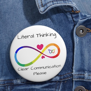 May include: A round white pinback button featuring the black text Literal Thinking and Clear Communication Please. In the center is a rainbow-colored infinity symbol with a pink heart and a small multicolored butterfly icon.