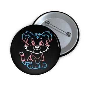 May include: A black button featuring a cartoon tiger cub design with pink and blue accents, representing the transgender pride flag. The button is round and has a pin on the back. A second button is visible in the background.