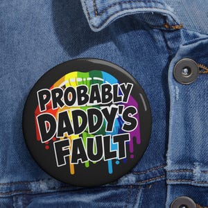Op de afbeelding: Een ronde button met een zwarte achtergrond en een afbeelding van regenboogkleurige, druipende lippen. De tekst PROBABLY DADDY'S FAULT staat in dikke, witte hoofdletters in het midden van het ontwerp, bevestigd op een blauwe spijkerstof.
