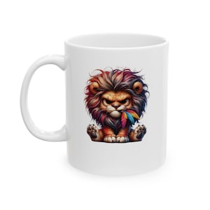 Puede incluir: Taza de cerámica blanca con un león de dibujos animados colorido con una pluma en la boca. El león tiene una expresión feroz y está sentado sobre sus cuartos traseros.