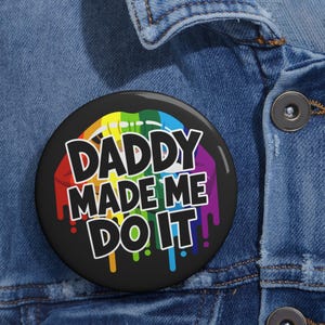 May include: A round pinback button featuring a black background with a graphic of rainbow-colored lips dripping paint. The text DADDY MADE ME DO IT is printed in bold white capital letters across the center of the colorful lip design.