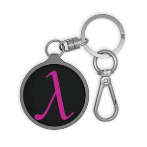 May include: A circular keychain featuring a black background with a bright pink Greek lowercase lambda symbol centered on the front. The keychain is attached to a silver-toned metal split ring and a lobster claw clasp for secure attachment.
