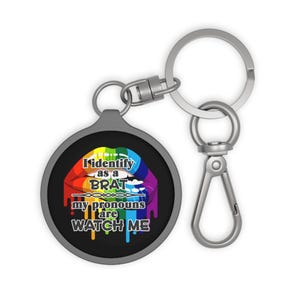 May include: A round metal keychain featuring a graphic of rainbow-colored lips with a dripping effect. The text reads: I identify as a BRAT my pronouns are WATCH ME. The design is set against a black background, attached to a silver split ring.