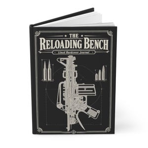 May include: A black hardcover journal titled The Reloading Bench with a white line art illustration of a rifle and various ammunition cartridges. The cover features technical blueprint-style graphics and text, with white pages visible at the top.