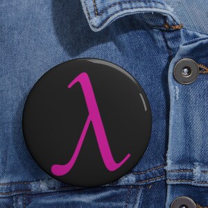 May include: A round pinback button featuring a solid black background with a bright magenta Greek lowercase lambda symbol centred on the surface. The button has a smooth, glossy finish and is attached to a blue denim fabric material.