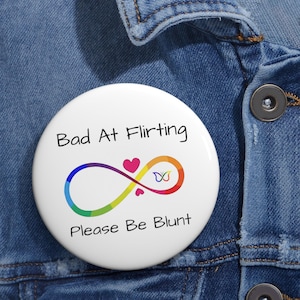 May include: A round white pin-back button featuring the black text Bad At Flirting and Please Be Blunt. In the centre is a rainbow-coloured infinity symbol with a small pink heart and a tiny multicoloured butterfly icon positioned above the loop.