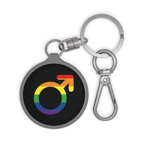May include: A round keychain with a black background featuring a rainbow-colored male symbol. The keychain has a silver-colored ring and clasp for attaching to keys or bags. The design is a symbol of LGBTQ+ pride.