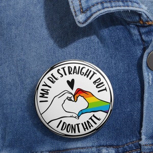 May include: A white button with black text and a rainbow heart design. The text reads "I may be straight but I don't hate".