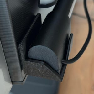 May include: A black charging cable and a black cylindrical object are held in place by a black, textured holder attached to a grey and white surface. The holder has a geometric design.