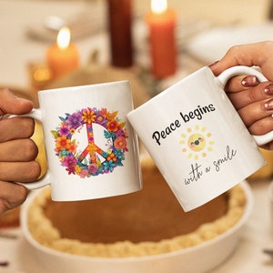 May include: Two white ceramic mugs with different designs. One mug has a peace sign surrounded by colorful flowers. The other mug has the text "Peace begins with a smile" and a yellow sun with a smiling face.