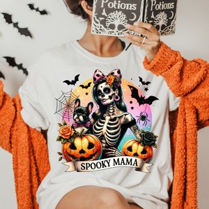 以下が含まれることがあります： ピンクのリボンをつけた骸骨の女性がフレンチブルドッグを抱いているカラフルなグラフィックがプリントされた白いTシャツ。グラフィックには、カボチャ、クモの巣、クモも含まれています。グラフィックの下には「Spooky Mama」という文字が書かれたバナーがプリントされています。