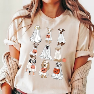 May include: Beige t-shirt featuring a Halloween-themed design. The graphic showcases various dog breeds dressed as ghosts, each holding a jack-o'-lantern. A small orange pumpkin is also included in the design. The shirt is a casual, everyday wear item.