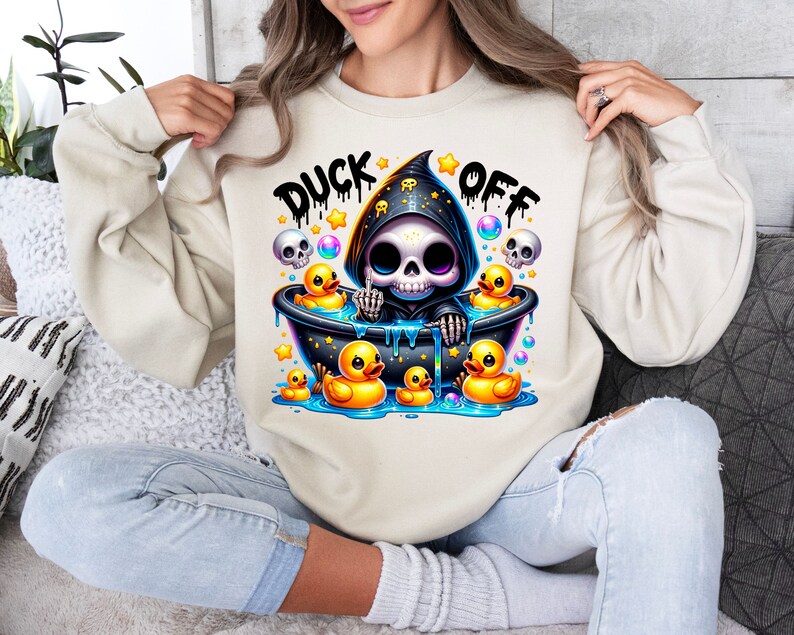 Może przedstawiać: Kremowa bluza z nadrukiem "Duck Off" z przodu. Tekst jest czarny i ma efekt kropel. Szkielet z kresk&oacute;wki w czarnej szacie z kapturem siedzi w wannie z gumowymi kaczkami. Szkielet trzyma gumową kaczkę w ręce.