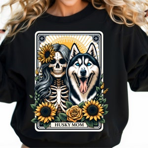 Puede incluir: Sudadera negra con un dise&ntilde;o gr&aacute;fico de una mujer esqueleto con girasoles y un perro husky. El texto "HUSKY MOM" est&aacute; impreso debajo de la imagen.