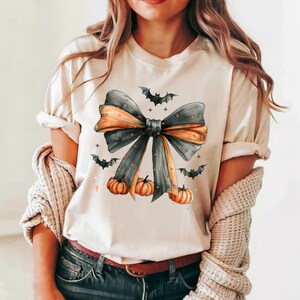 May include: A cream-colored t-shirt with a black and orange bow with gold glitter accents. The bow is surrounded by small black bats and orange pumpkins.