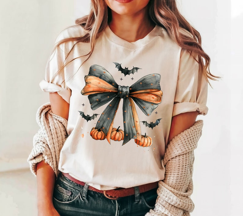 May include: A cream-colored t-shirt with a black and orange bow with gold glitter accents. The bow is surrounded by small black bats and orange pumpkins.